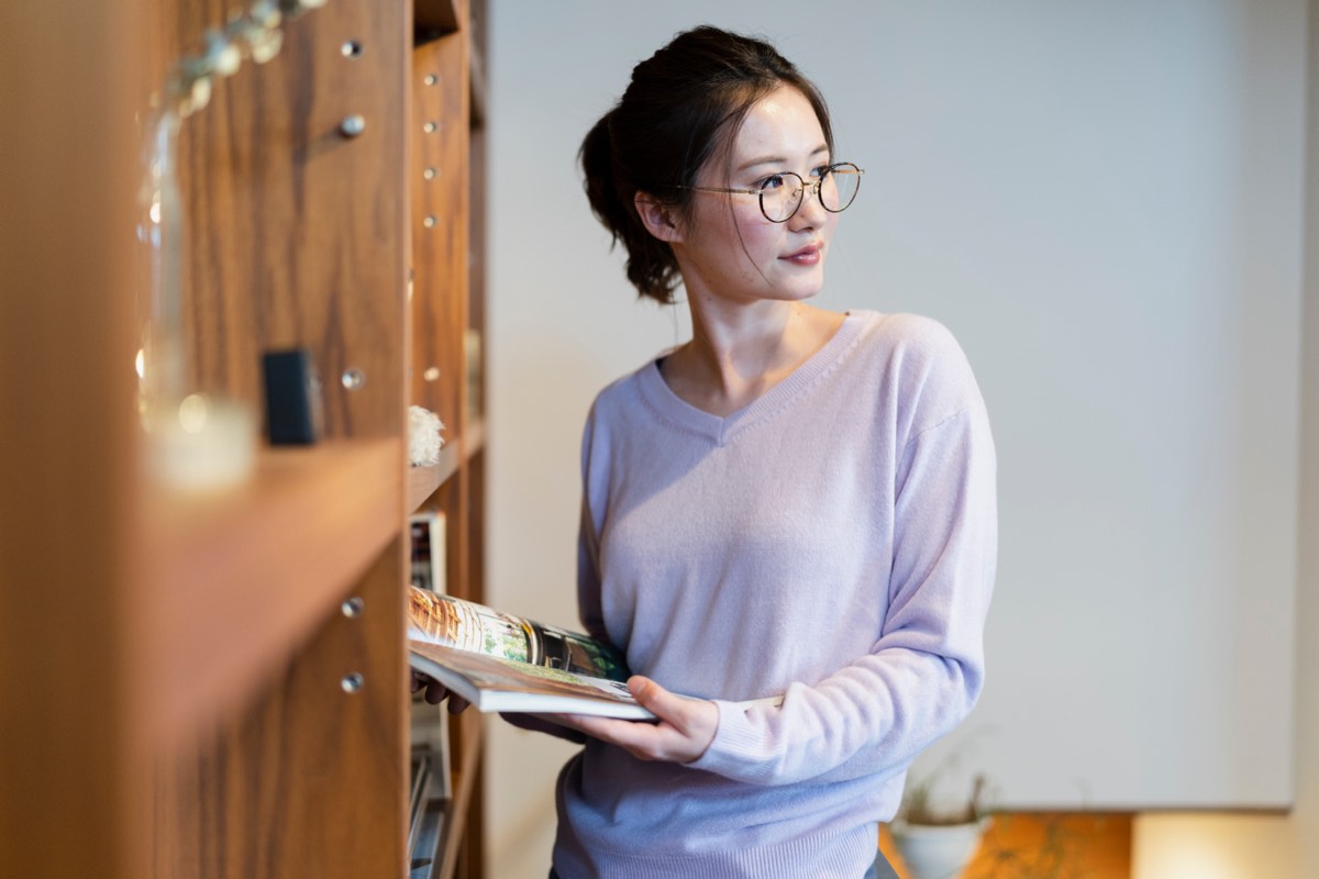 本棚の前で本を開く女性。読む順番を選ぶイメージ