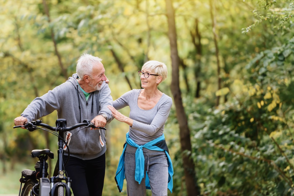 自転車を押して公園を歩く笑顔のシニア夫婦。健康と趣味が資産になる60代以降